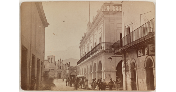 A sepia-toned photograph of a street scene with tall buildings on either side, people and carriages in the street, mountains in the distance.