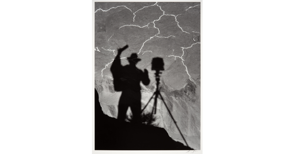 A black-and-white photograph of the shadow of a person and tripod with camera cast from above onto the landscape below.