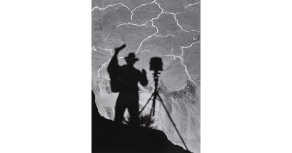 A black-and-white photograph of the shadow of a person and tripod with camera cast from above onto the landscape below.