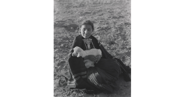 A black-and-white photograph of an Indigenous girl sitting on the ground while holding a lamb.