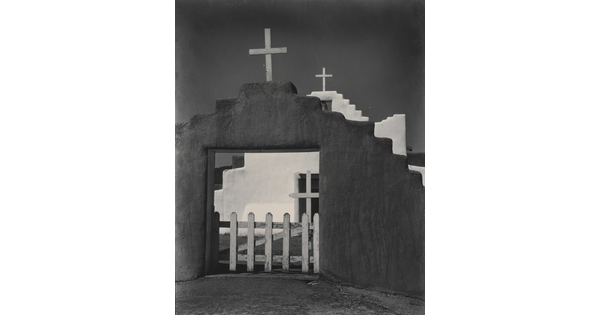 A black-and-white photograph of a white adobe church seen through the entrance of the surrounding adobe wall topped with a wooden cross.