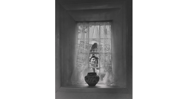 A black-and-white photograph of a sheer curtained window with a Native American-style pot on the sill as a White woman cleans the window from outside.