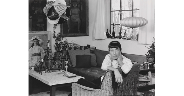 A black-and-white photograph of a White woman sitting sideways in a chair, facing the camera, in a room full of art, sculpture, bottles, candlesticks, etc.