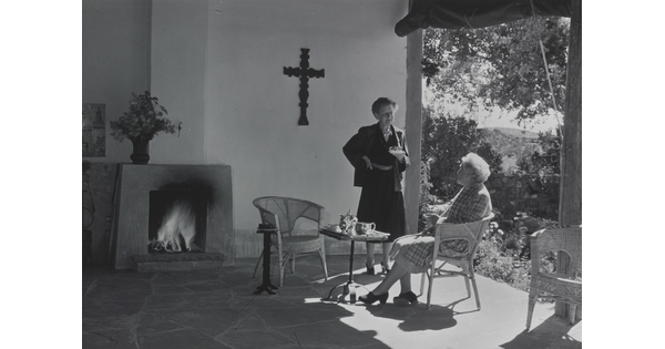 A black-and-white photograph of two White women, one seated and one standing, talking on a covered patio.