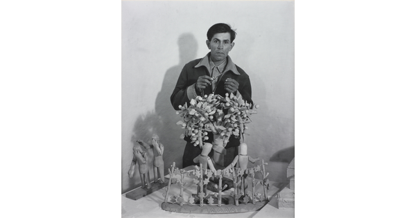 A black-and-white photograph of a medium-skinned man standing behind a wood sculpture of trees and human figures.