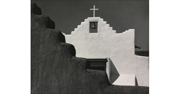 A black-and-white photograph of a terraced adobe bell tower with a cross on top, partially hidden by a terraced adobe wall.