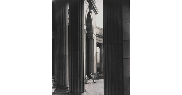 A black-and-white photograph of a person sitting on a bench reading a newspaper as seen from between large fluted columns.