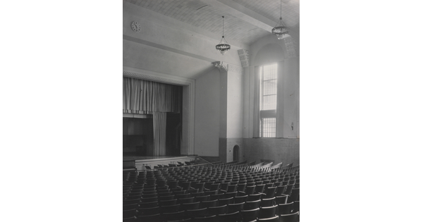 A black-and-white photograph of an empty auditorium showing rows of seats, a stage, and a tall window.