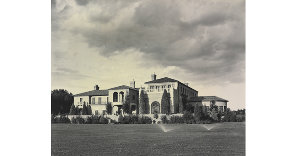 A black-and-white photograph of a large multi-story mansion with a mission-style roof and a large grass yard with sprinklers on.