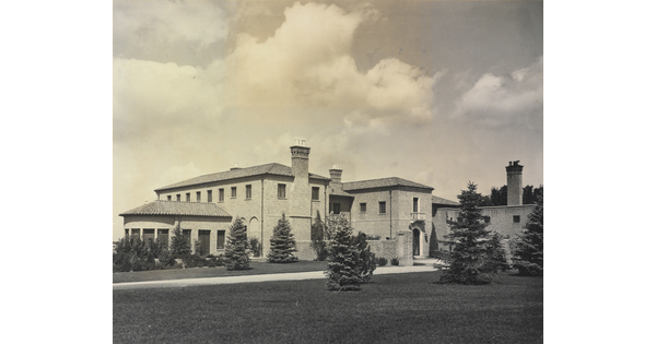A black-and-white photograph of a large multi-story mansion with a mission-style roof and a large grass yard with evergreens.