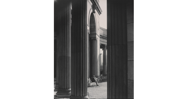 A black-and-white photograph of a person sitting on a bench reading a newspaper as seen from between large fluted columns.