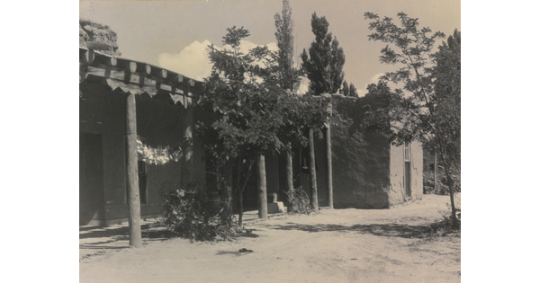 A black-and-white photograph of the exterior of a single-story adobe house and covered porch surrounded by trees and a dirt yard.
