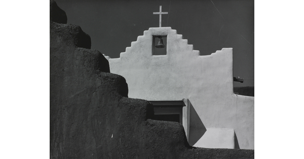 A black-and-white photograph of a terraced adobe bell tower with a cross on top, partially hidden by a terraced adobe wall.