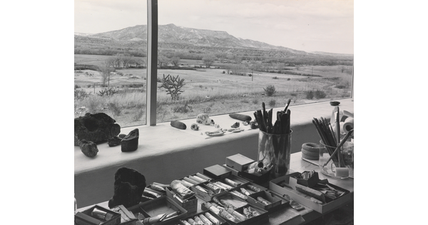 A black-and-white photograph of paints, brushes, and other art supplies on a wide windowsill that overlooks a mountain landscape.