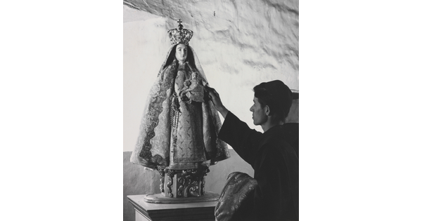 A black-and-white photograph of a friar touching a statue of the Virgin Mary dressed as queen of heaven holding the infant Jesus.