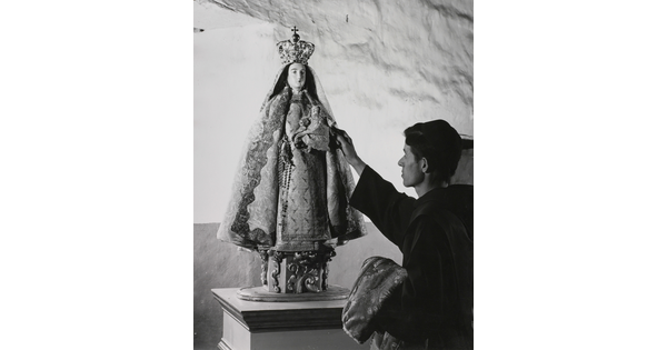 A black-and-white photograph of a friar touching a statue of the Virgin Mary dressed as queen of heaven holding the infant Jesus.