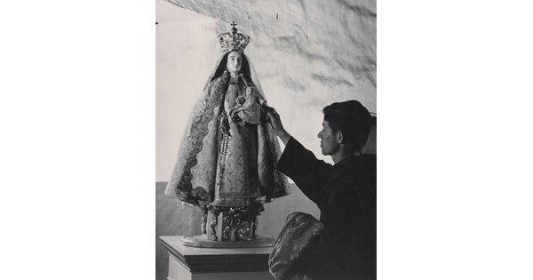 A black-and-white photograph of a friar touching a statue of the Virgin Mary dressed as queen of heaven holding the infant Jesus.