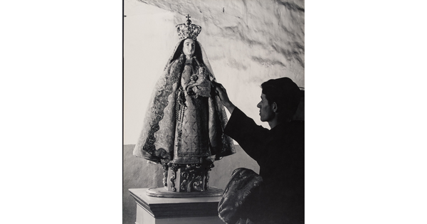 A black-and-white photograph of a friar touching a statue of the Virgin Mary dressed as queen of heaven holding the infant Jesus.