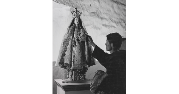 A black-and-white photograph of a friar touching a statue of the Virgin Mary dressed as queen of heaven holding the infant Jesus.