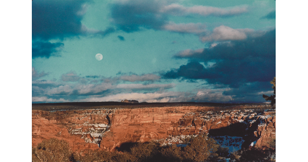 A color photograph of an almost-full moon in a cloudy blue sky over red, rocky cliffs with snow in the crevices.