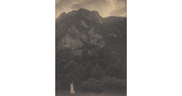 A black-and-white photograph of a woman in a white dress standing at the base of a rocky mountain covered with trees.