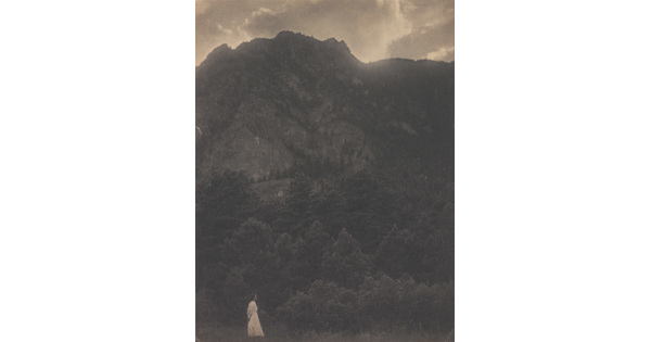 A black-and-white photograph of a woman in a white dress standing at the base of a rocky mountain covered with trees.