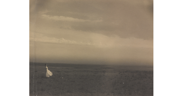 A black-and-white photograph of a woman in a white dress standing in a large meadow.