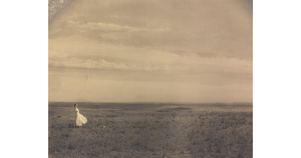 A black-and-white photograph of a woman in a white dress standing in a large meadow.