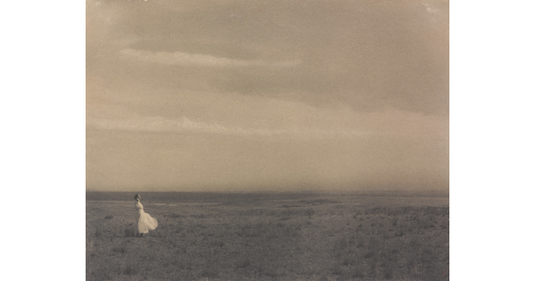 A black-and-white photograph of a woman in a white dress standing in a large meadow.