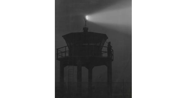A black-and-white photograph of a person standing in silhouette atop a tower with a bright spotlight shining from the top.