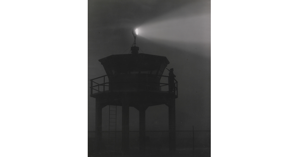A black-and-white photograph of a person standing in silhouette atop a tower with a bright spotlight shining from the top.