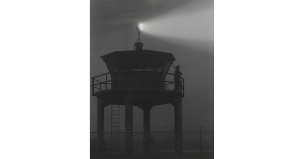 A black-and-white photograph of a person standing in silhouette atop a tower with a bright spotlight shining from the top.