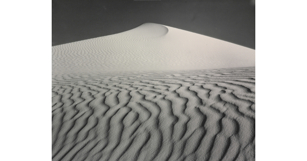 A sepia-toned photograph of a deeply-rippled sand dune rising to a point into the sky.