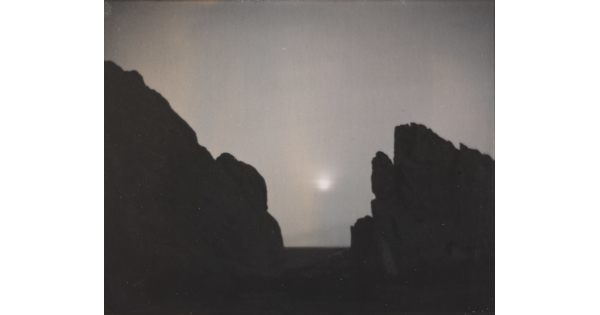 A black-and-white photograph of a moon shining through a foggy sky from between the V-shaped silhouette of large rocks.