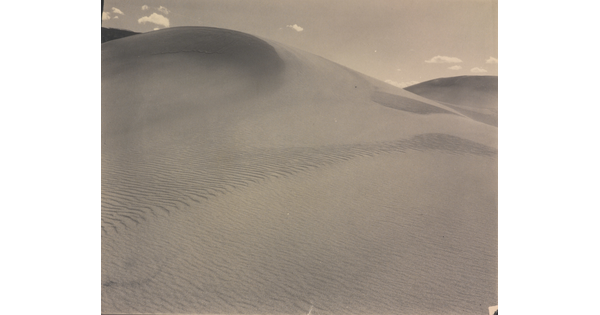 A sepia-toned photograph of a rippling sand dune rising up into a sky dotted with a few small clouds.