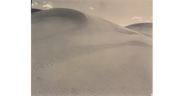 A sepia-toned photograph of a rippling sand dune rising up into a sky dotted with a few small clouds.