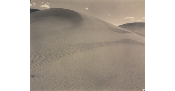 A sepia-toned photograph of a rippling sand dune rising up into a sky dotted with a few small clouds.