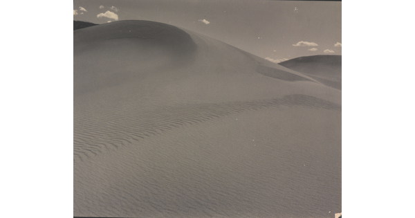 A sepia-toned photograph of a rippling sand dune rising up into a sky dotted with a few small clouds.