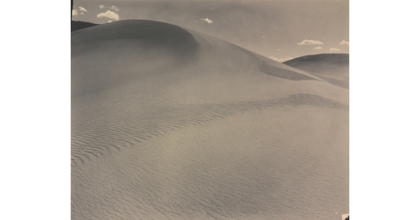 A sepia-toned photograph of a rippling sand dune rising up into a sky dotted with a few small clouds.