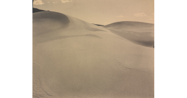 A sepia-toned photograph of a rippling sand dune rising up into a sky dotted with a few small clouds.