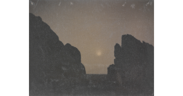 A black-and-white photograph of two rock formations in silhouette with a hazy moon in between them.