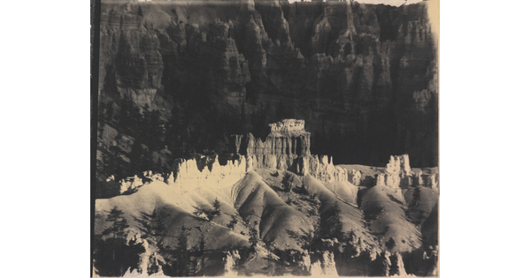 A black-and-white photograph of a landscape with hoodoos, or vertical spires of rock, on top of sand dune shapes with cliffs in the background.