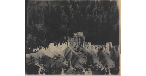 A black-and-white photograph of a landscape with hoodoos, or vertical spires of rock, on top of sand dune shapes with cliffs in the background.