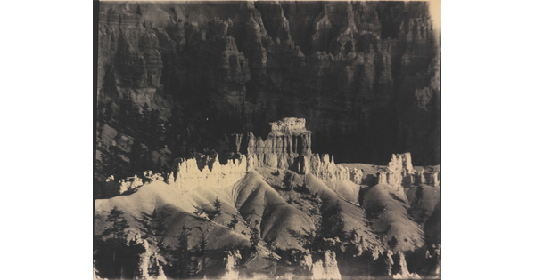 A black-and-white photograph of a landscape with hoodoos, or vertical spires of rock, on top of sand dune shapes with cliffs in the background.