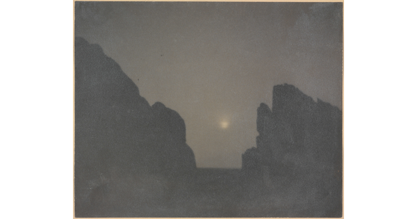 A black-and-white photograph of two rock formations in silhouette with a hazy moon in between them.