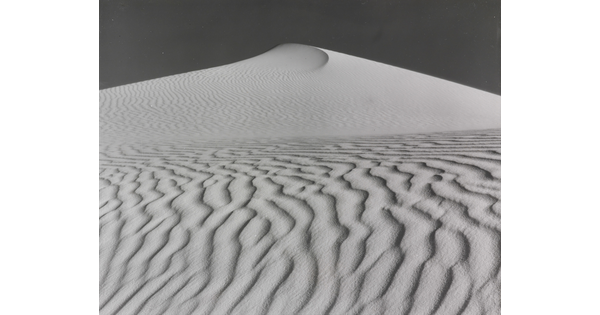 A sepia-toned photograph of a deeply-rippled sand dune rising to a point into the sky.