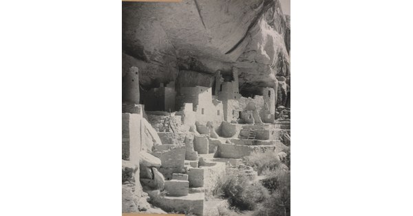 A black-and-white photograph of an abandoned, crumbling rock city built into the side of a desert cliff.