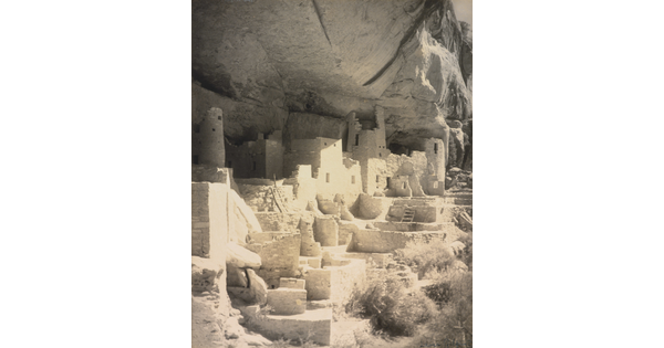 A black-and-white photograph of an abandoned, crumbling rock city built into the side of a desert cliff.