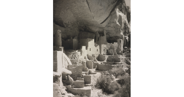 A black-and-white photograph of an abandoned, crumbling rock city built into the side of a desert cliff.
