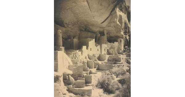 A black-and-white photograph of an abandoned, crumbling rock city built into the side of a desert cliff.